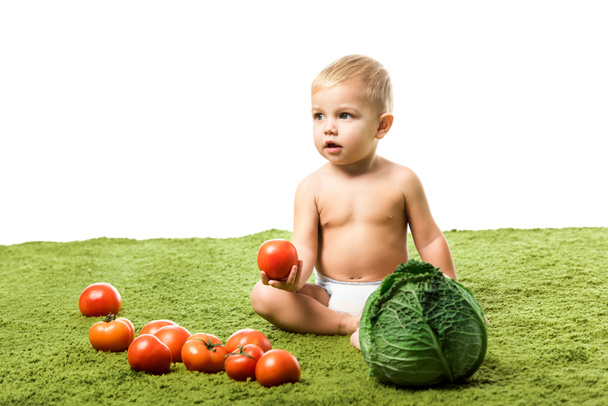 schattig peuter jongen houdt van tomaat en zittend op het groene tapijt met groenten geïsoleerd op wit - Foto, afbeelding