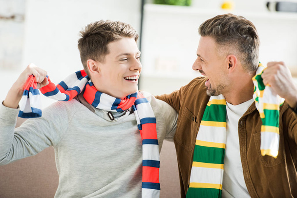 happy father and teen son in scarfs watching sport match at home, looking at each other - Foto, immagini