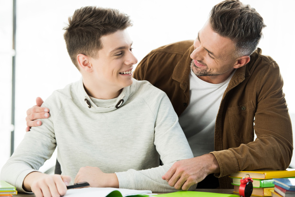 smiling father hugging teen son while he doing homework, looking at each other - Photo, image