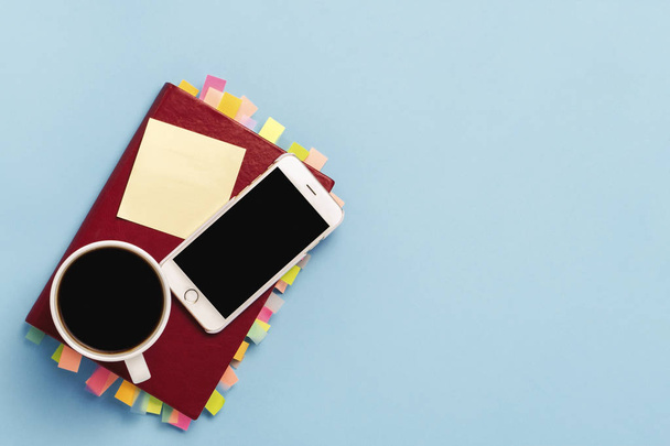 Red diary with stickers on the pages, a cup with black coffee, white phone, blue background. Concept of a successful business, proper planning, time management. Flat lay, top view. - Foto, Bild