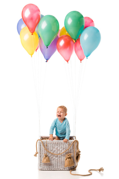 μικρό παιδί αγόρι κλαίει και στέκεται σε ψάθινο καλάθι με πολύχρωμα μπαλόνια που απομονώνονται σε λευκό - Φωτογραφία, εικόνα