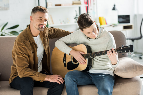teen son playing acoustic guitar for father at home - Фото, изображение