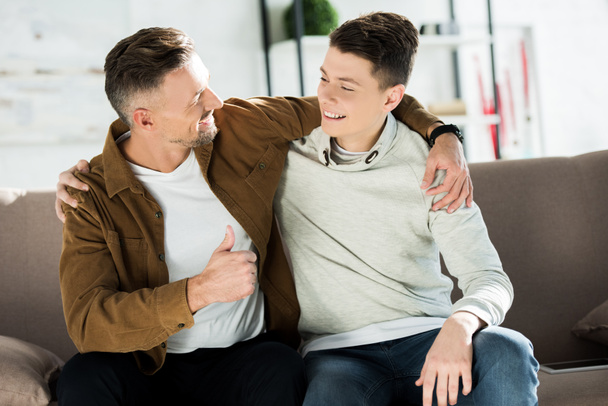 smiling father and teen son hugging on sofa at home and looking at each other - Foto, Imagen