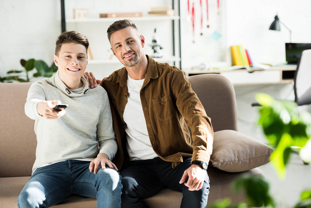 hymyilevä isä ja teini poika katsomassa televisiota yhdessä sohvalla olohuoneessa
 - Valokuva, kuva