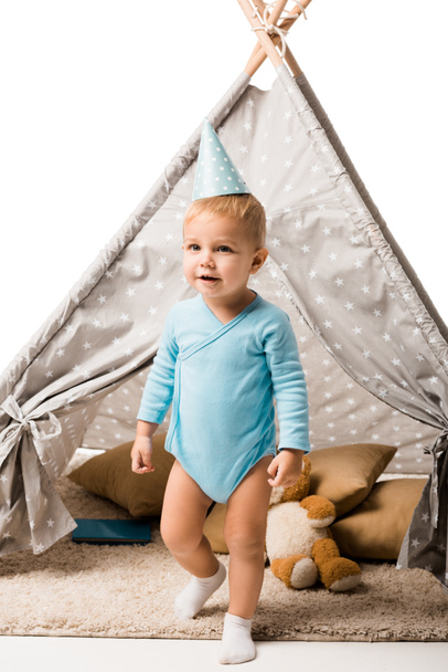 toddler boy in party hat standing in front of wigwam with pillows and teddy bear isolated on white - Photo, Image
