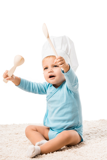toddler boy in chefs hat sitting on carpet and holding two big wooden spoons isolated on white - Foto, immagini