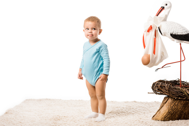 cute toddler boy in blue bodysuit standing on carpet near big decorative stork in nest isolated on white - Photo, Image