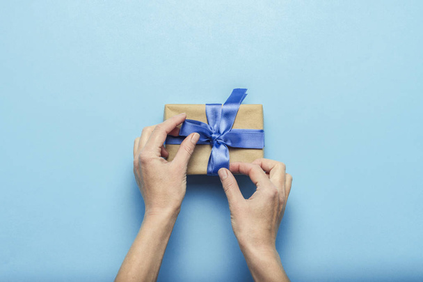 Female hands tying a bow on a gift box with a blue ribbon on a blue background. Concept of a gift for the holidays, birthday, Christmas, wedding. Flat lay, top view. - Foto, imagen