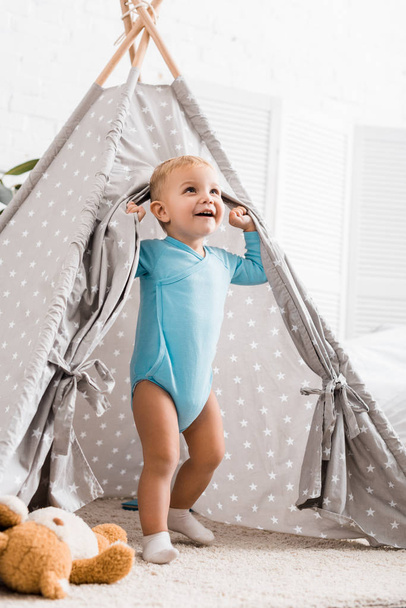 söpö lapsi poika sininen kehonpuku hymyilee ja seisoo harmaa vauva wigwam lastenhuoneessa
 - Valokuva, kuva