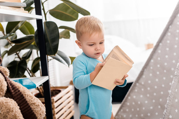 cute toddler boy in blue bodysuit holding book in bedroom - Photo, Image