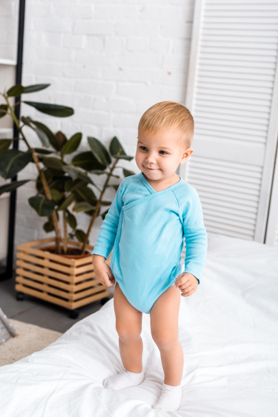 smiling baby in blue bodysuit standing on bed in nursery room - Foto, imagen