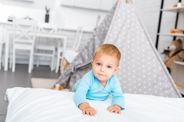 cute toddler boy standing near bed, smiling and looking at camera in apartment - Photo, Image
