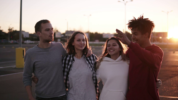 Group of mixed race friends walking embracing and talking together in city. They are two girls and two boys in their twenties, friendship and lifestyle concepts, autumn clothing. Sun strongly shining - Footage, Video