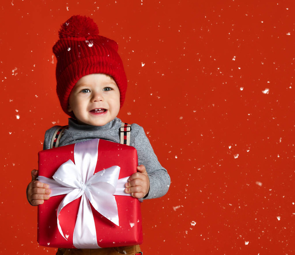Portrait of a little boy in a red hat with a pompon. holding a large gift box with a white bow.  - Photo, image