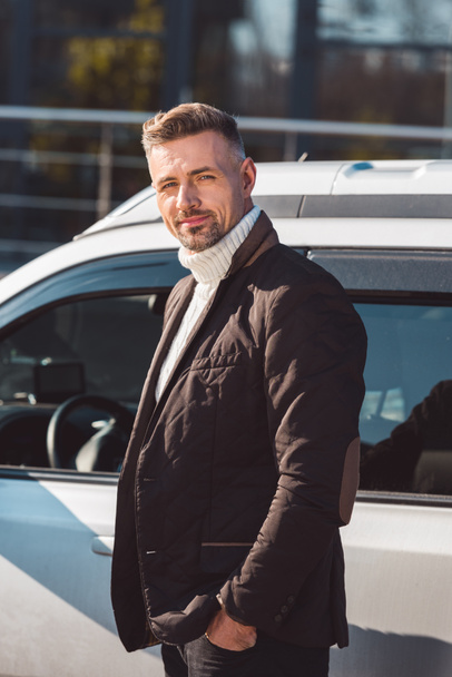 Cheerful man standing and smiling near car with hands in pockets - Photo, Image