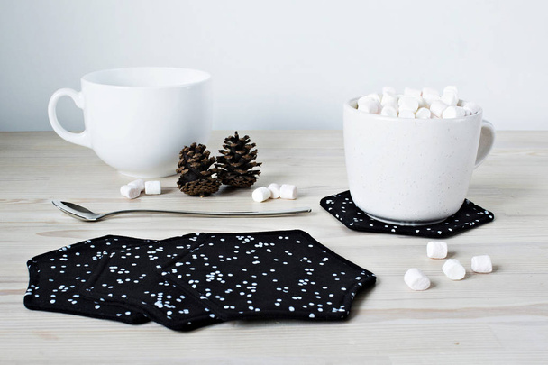 Black fabric coasters, white cups, pine cones, spoon and marshmallows on the table - Valokuva, kuva