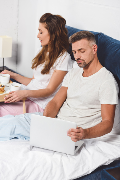 handsome husband using laptop while attractive wife having breakfast in bed  - Фото, изображение