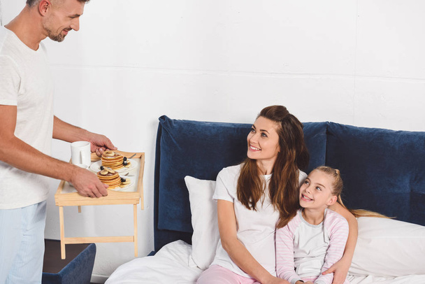 father holding wooden tray with pancakes while daughter and wife smiling and sitting in bed - Φωτογραφία, εικόνα