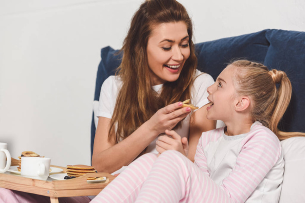 smiling mother feeding daughter in bed during breakfast - Фото, зображення