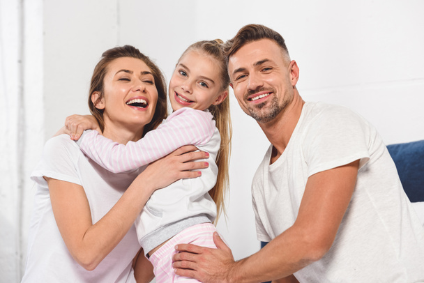 cute daughter hugging beautiful smiling mother in bed while father looking at camera - Photo, image
