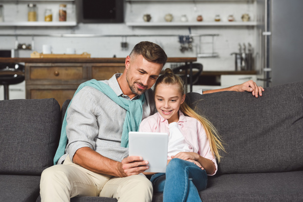 father and daughter sitting on sofa and using digital tablet - Фото, изображение