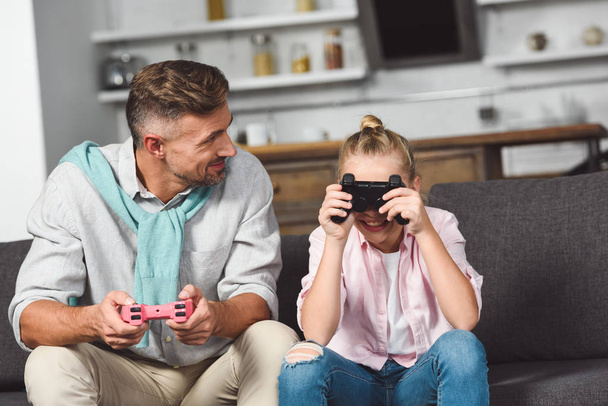 father looking at daughter while child hiding eyes behind joystick - Photo, Image