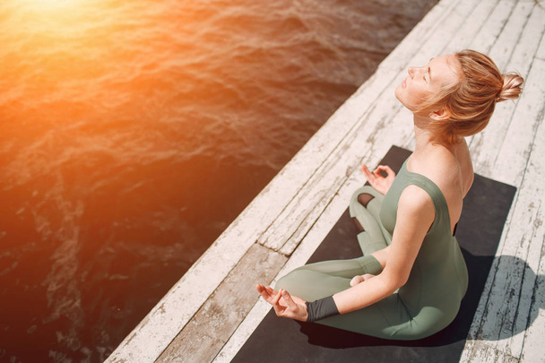 Young attractive woman practicing yoga at lake shore. Sun flare. Toned. Copy space. - Photo, Image
