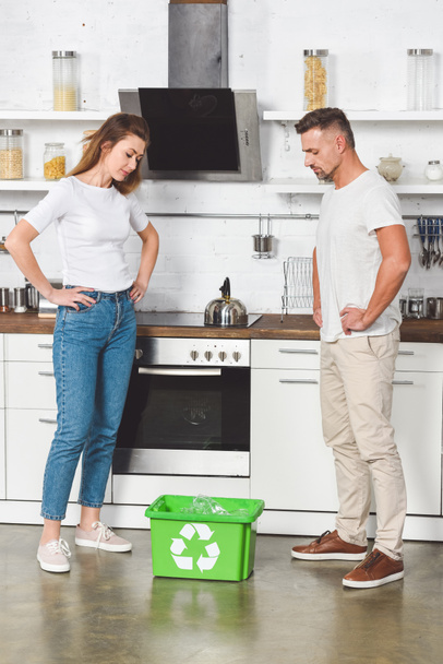 pareja adulta con manos en montones de pie en la cocina y mirando la caja de reciclaje verde
 - Foto, imagen