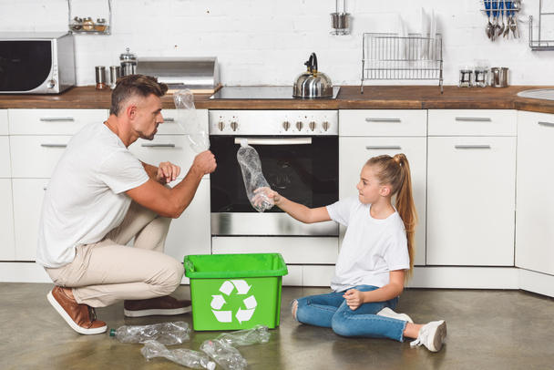 Ojciec i córka siedzi na podłodze w kuchni i wprowadzenie puste butelki plastikowe pudełko z recyklingu znak - Zdjęcie, obraz