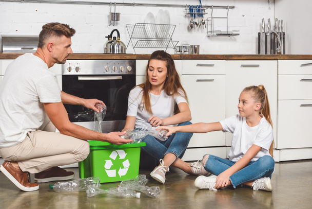 familia sentada en el suelo en la cocina y poniendo botellas de plástico vacías en la caja con signo de reciclaje
 - Foto, Imagen