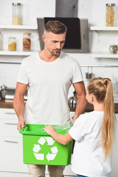 Baba ve kızı mutfakta yeşil geri dönüşüm kutusunu boş plastik şişe koyarak - Fotoğraf, Görsel