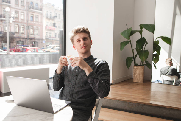Przystojny młody mężczyzna ubrany w koszulę siedzi w światło kawiarni, z filiżanką kawy w jego ręce, laptopa na stole. Szczęśliwy student pije kawę w kawiarni i używa laptopa. - Zdjęcie, obraz