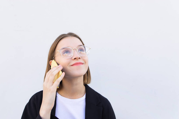 Ευτυχισμένος φοιτητής κορίτσι με τα γυαλιά στέκεται σε ένα λευκό φόντο και συνομιλίες πάνω από το τηλέφωνο. Πορτρέτο του ένα χαμογελαστό γυναίκα επιχείρησης καλώντας στο τηλέφωνο. Copyspace - Φωτογραφία, εικόνα