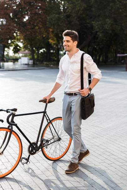 Happy businessman dressed in shirt walking with a bicycle outdoors at the city street, carrying a bag - Foto, afbeelding