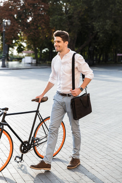 Handsome businessman dressed in shirt walking with a bicycle outdoors at the city street, carrying a bag - Photo, Image