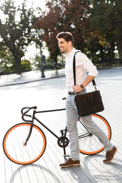 Smiling businessman dressed in shirt walking with a bicycle outdoors at the city street, carrying a bag - Photo, image