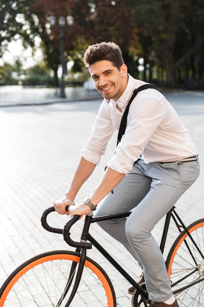 Cheerful businessman dressed in shirt riding on a bicycle outdoors at the city street, carrying a bag - 写真・画像