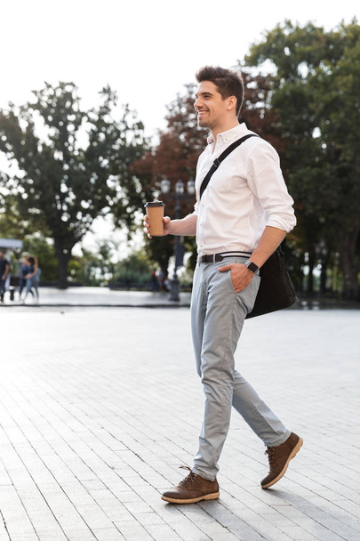 Confident businessman dressed in shirt walking outdoors at the city street, holding takeaway coffee - Фото, изображение