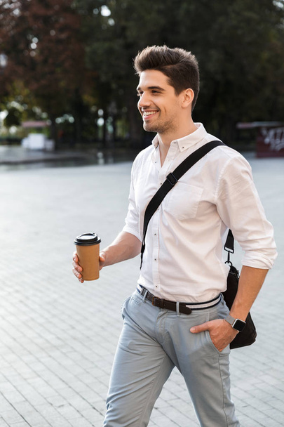 Positive businessman dressed in shirt walking outdoors at the city street, holding takeaway coffee - Foto, afbeelding