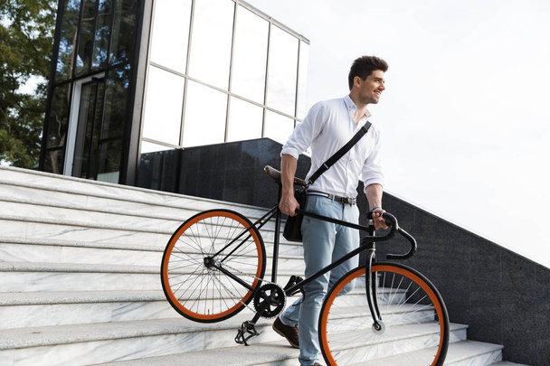 Happy businessman dressed in shirt carrying a bicycle outdoors at the city street, going downstairs - Fotoğraf, Görsel