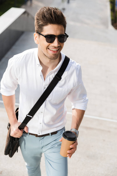 Happy businessman dressed in shirt walking upstairs outdoors at the city street, holding takeaway coffee - Fotó, kép