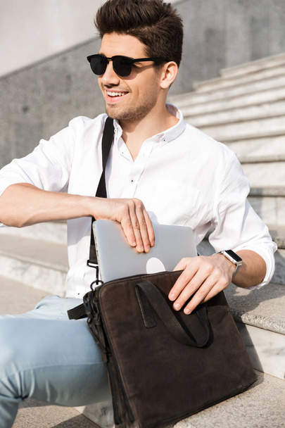 Photo of handsome man 30s wearing sunglasses, putting laptop into his bag while sitting on stairs on city street - Fotografie, Obrázek