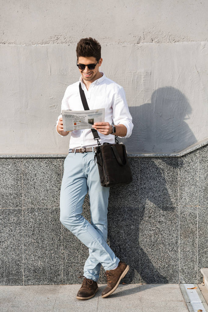 Photo of young man 30s wearing sunglasses reading newspaper while standing along wall outdoor - Foto, afbeelding