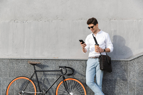 Photo of smiling man 30s wearing sunglasses, drinking takeaway coffee and using mobile phone, while standing with bicycle along wall outdoor - Foto, Bild