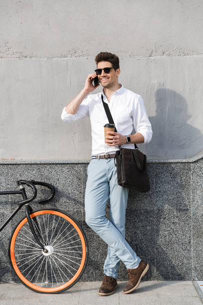 Photo of happy man 30s wearing sunglasses drinking takeaway coffee and talking on mobile phone while standing with bicycle along wall outdoor - Fotoğraf, Görsel