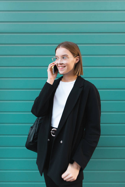 Mujer joven feliz en ropa elegante se encuentra en el fondo de una pared de color turquesa y habla por teléfono. Mujer de negocios sonriente sobre un fondo de color
. - Foto, Imagen