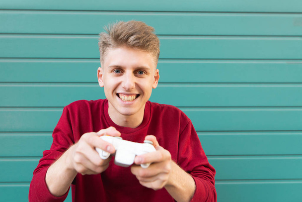 Happy gamer with a gamepad in his hands plays a console on a turquoise background and looks at the camera. Teen plays a joystick on a colored background. - Zdjęcie, obraz