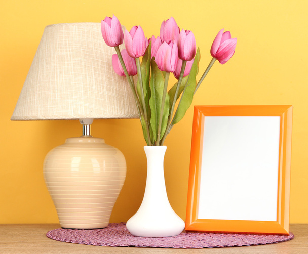Colorful photo frame, lamp and flowers on wooden table on yellow background - Foto, Imagem