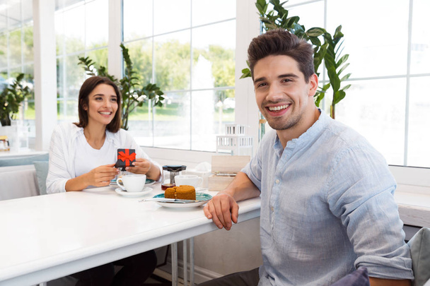 Image of amazing young loving couple sitting in cafe eat desserts and drinking tea. Man looking at camera while his girlfriend looking at her present. - Fotografie, Obrázek