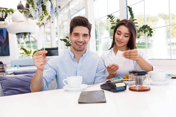Image of a young happy loving couple sitting in cafe holding credit card and check. - Foto, Imagem